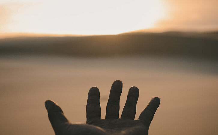 Eine Hand, die sich dem bei Sonnenuntergang warm schimmernden Horizont entgegenstreckt und Vergebung und Loslassen symbolisiert.